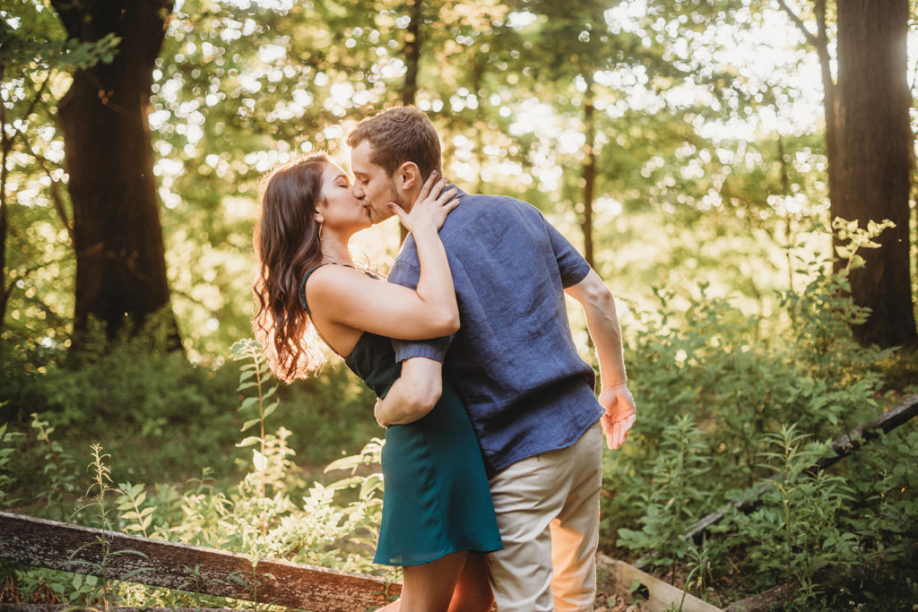 young man hugs woman with one arm and kisses her at sunset in woods at an indy park