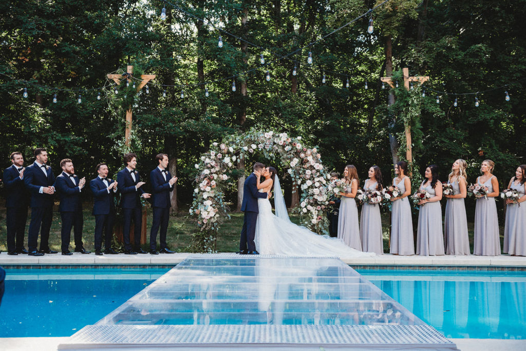bride and groom first kiss wideshot with pool visible and wedding party all watching