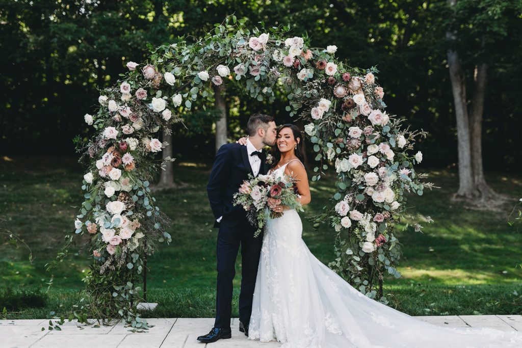 groom kisses bride in front of flower arch at their Carmel Backyard Wedding