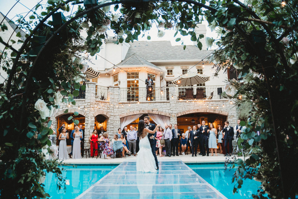 wide shot through flower arch of bride and groom dancing over a pool for their first dance at their Carmel Backyard Wedding