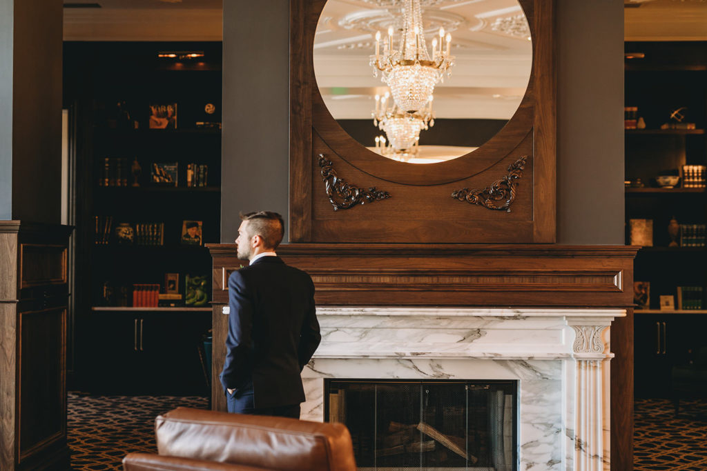 Groom stands in front of fireplace in Hotel Carmichael