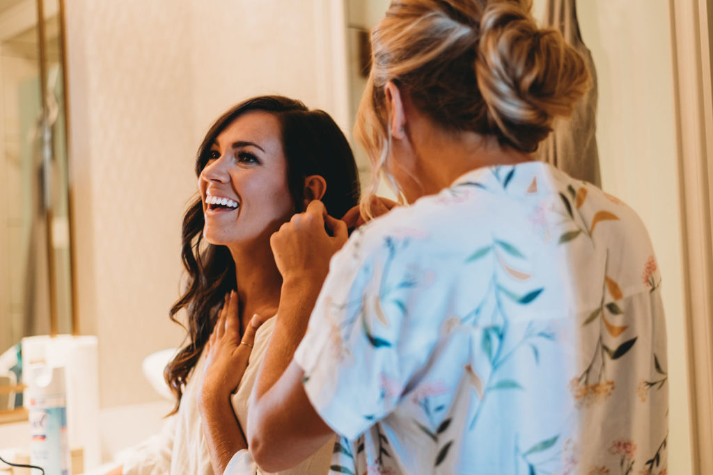beautiful young bride has earrings put on by bridesmaid