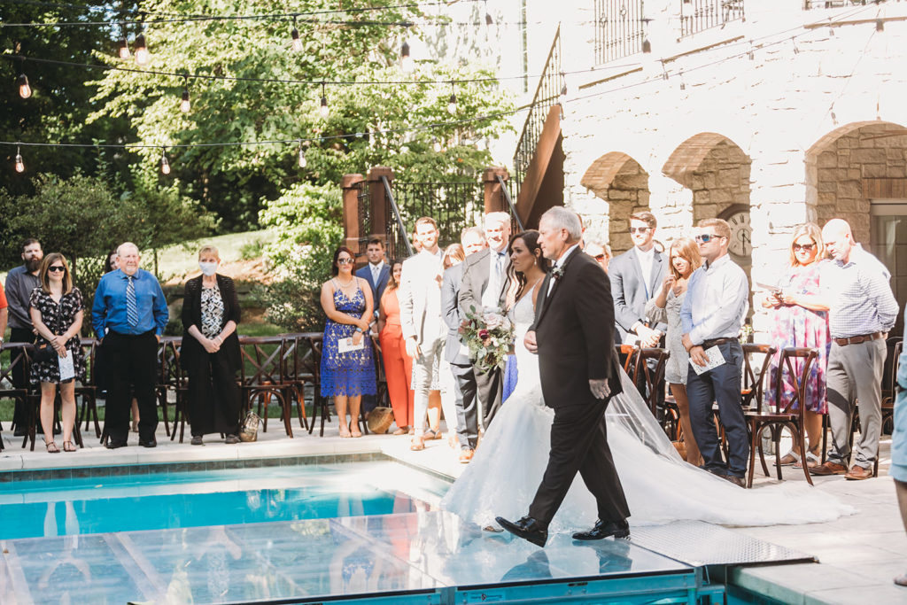 father of the bride walks the bride across a glass platform over a pool to her Carmel Backyard Wedding