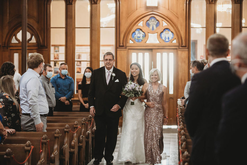bride walks down aisle of St. John the Evangelist Catholic Church with her mom and dad