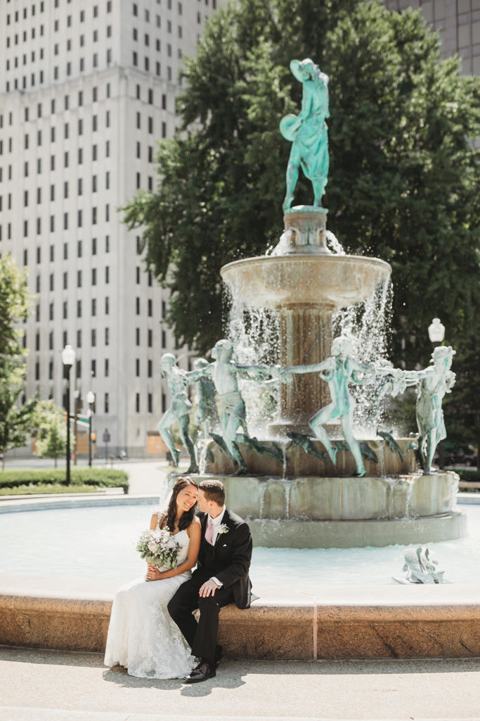 bride and groom sit in front of fountain in Indianapolis park before their Indiana Roof Ballroom wedding