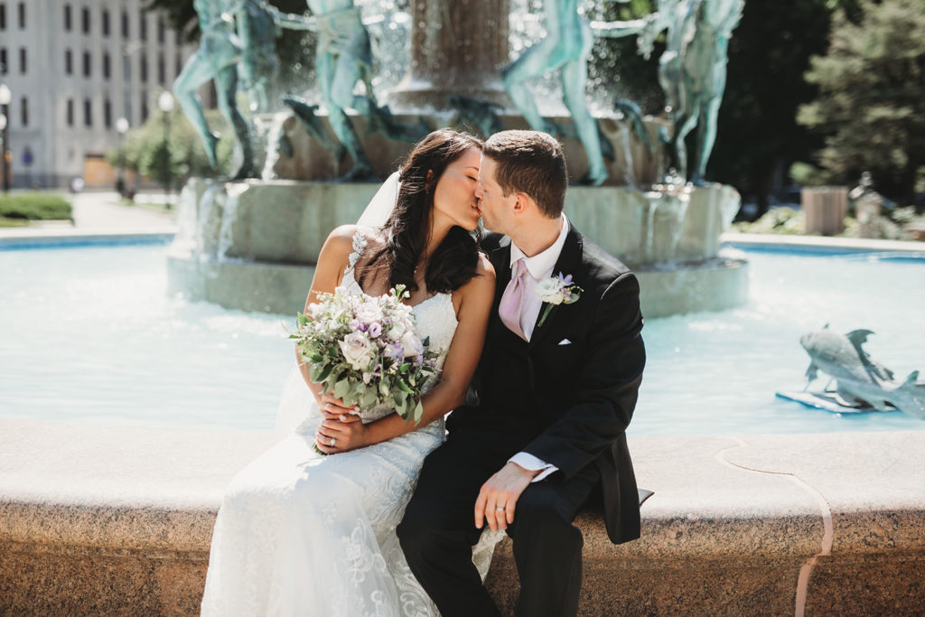 bride and groom kiss in front of fountain in Indianapolis park before their Indiana Roof Ballroom wedding