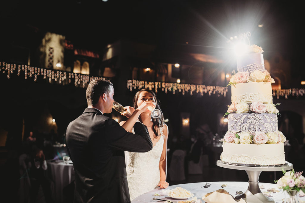 bride and groom stand next to 4 tier wedding cake and drink champagne together at their Indiana Roof Ballroom wedding