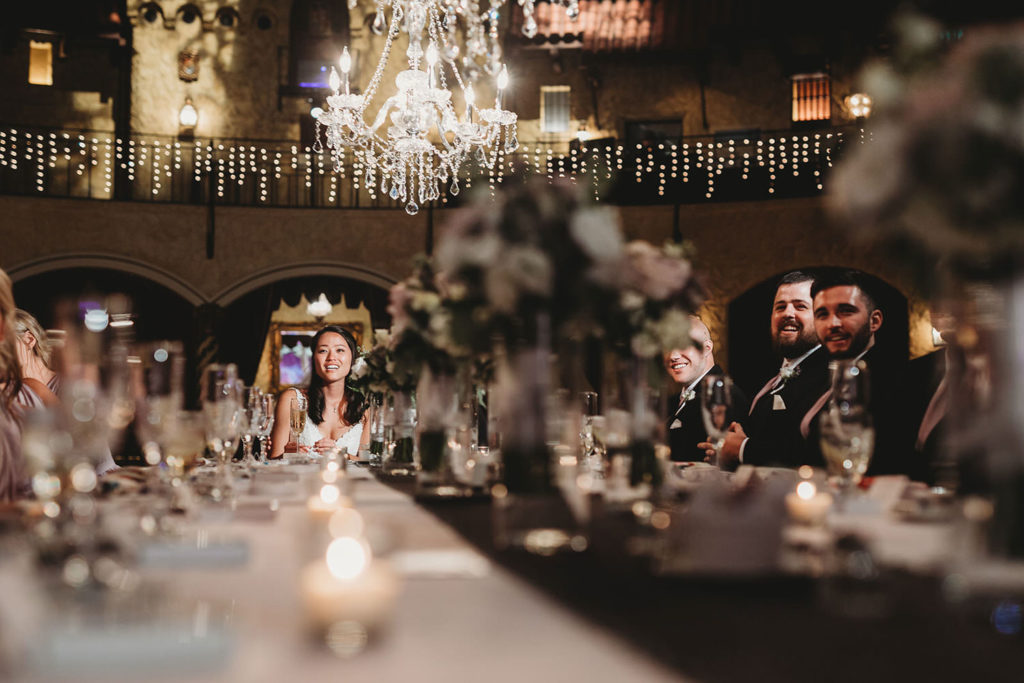 shot of bride watching toasts at her Indiana Roof Ballroom wedding with floral on long family style table acting as a leading line to her face