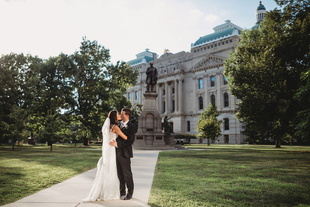 bride and groom hug and kiss in front of statue in front of indiana statehouse during their Indiana Roof Ballroom wedding