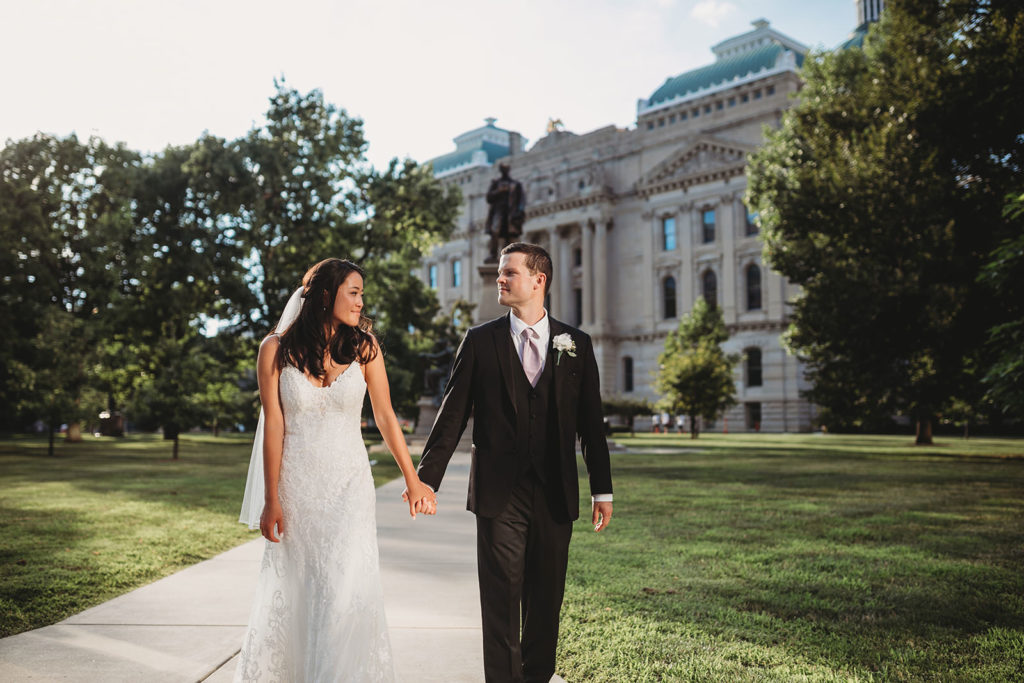 bride and groom hold hands and walk away from the Indiana Statehouse during their Indiana Roof Ballroom wedding
