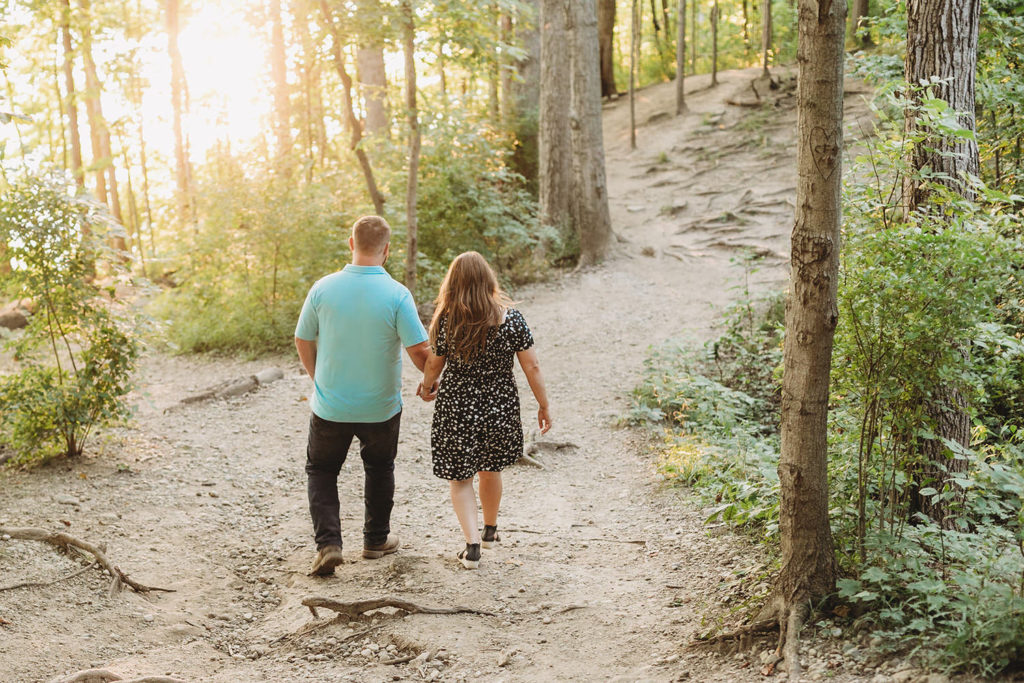 couple holds hands and walks through woods toward the setting sun during their Indy Reservoir Engagement