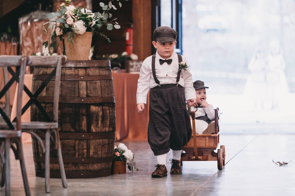 little boys dressed like Newsies walk down the aisle, one pulling the other in a wagon at a 3 Fat Labs wedding