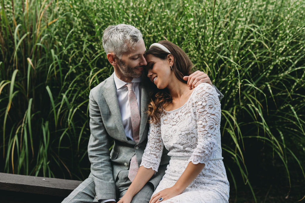 bride and groom snuggle on wooden bench in front of tall grass at their traders point creamery wedding