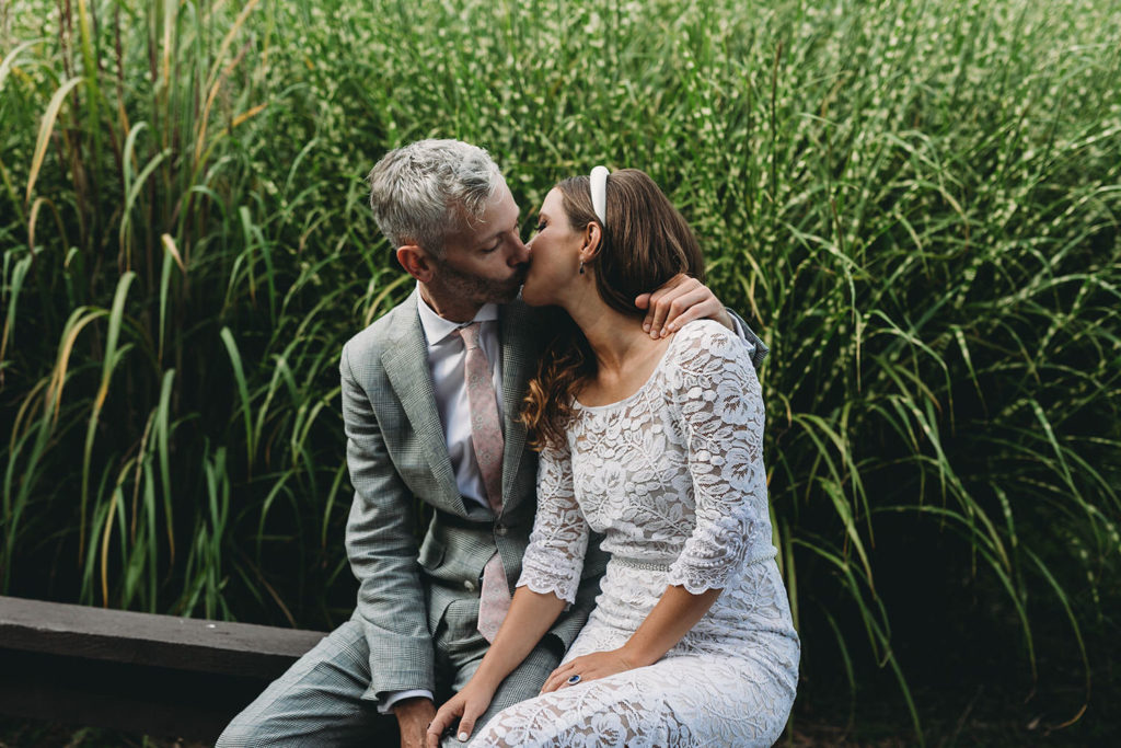 bride and groom kiss on wooden bench in front of tall grass at their traders point creamery wedding