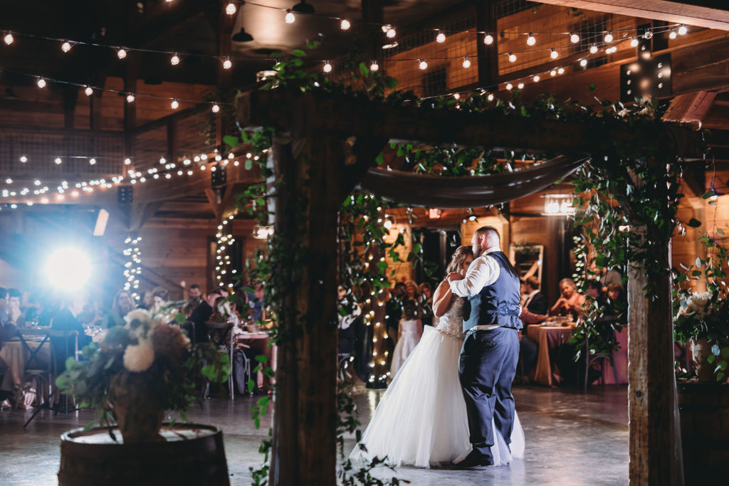 bride and groom framed by wooden arch covered in hanging greenery during their first dance at their 3 Fat Labs wedding