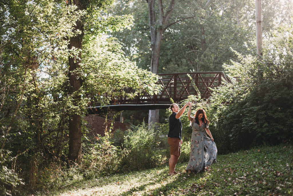 man dances with woman in green floral dress in front of bridge in woods during their Arbuckle Acres engagement session