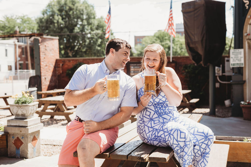 couple sit on picnic table with beers and laugh during their Rathskeller engagement session