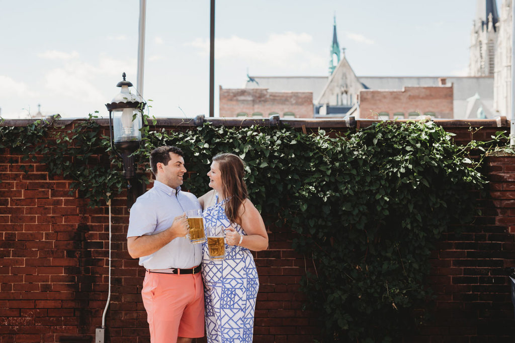 man and woman stand in front of hanging ivy with beer during their Rathskeller engagement session