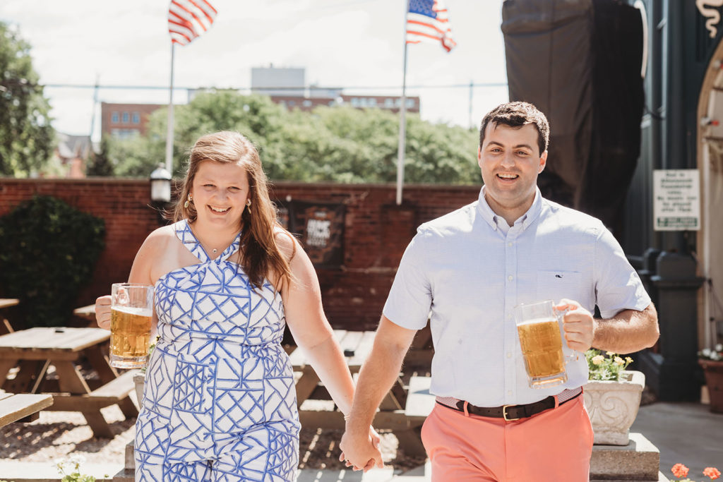 couple hold hands and hold beer while walking through biergarten during their Rathskeller engagement session