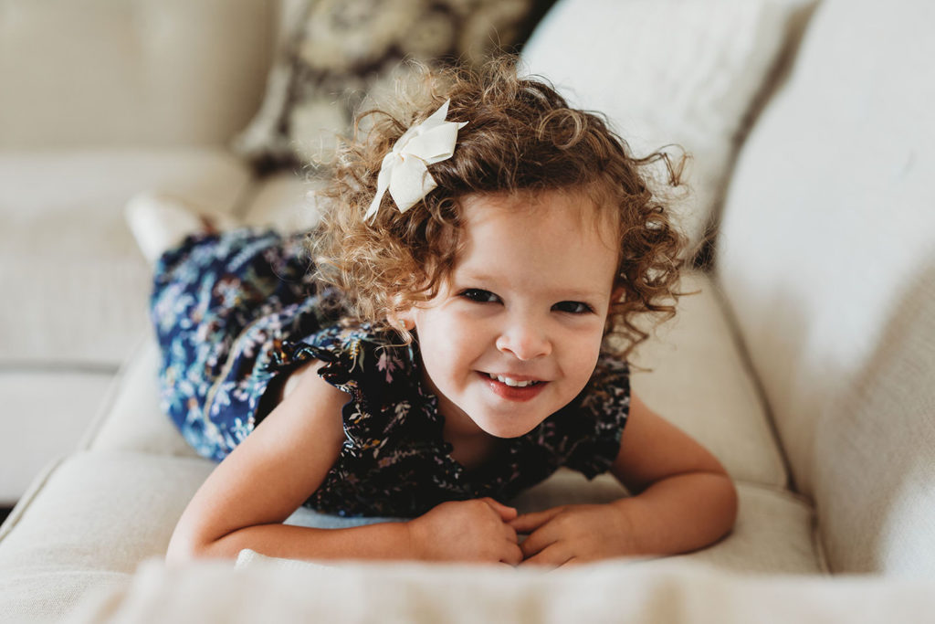 girl with curly hair and blue dress on white couch smiling in this parker city family photography