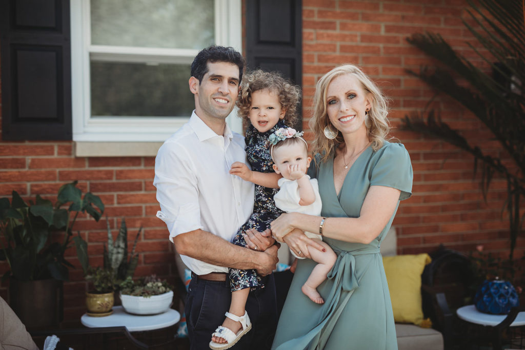 blonde mom and dark haired dad in front of brick house holding daughters for this farmland family photography