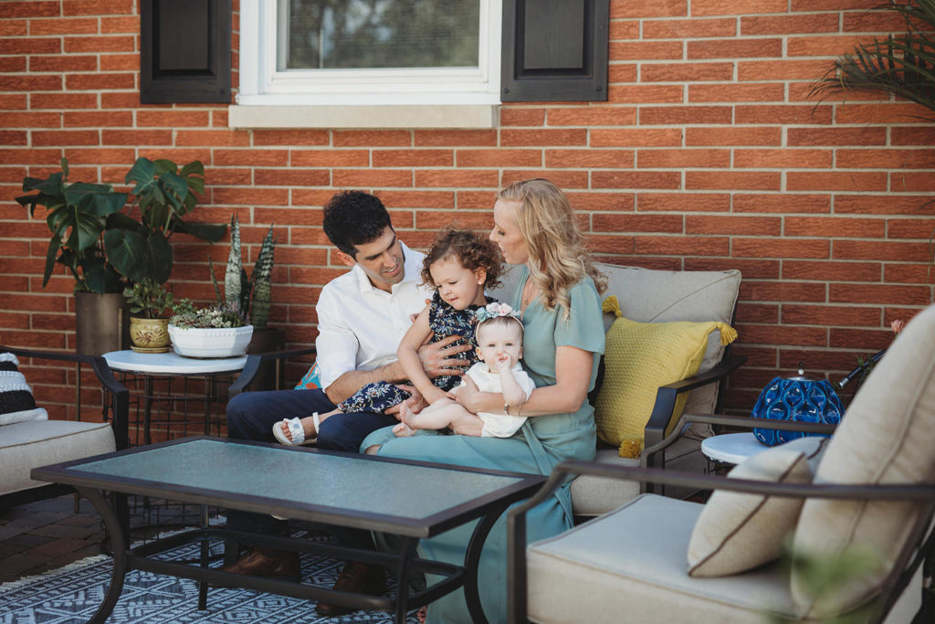 dad and mom hug kids on patio furniture in front of brick house in these parker city family portraits