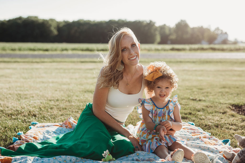 blonde mother in green skirt and white tank top sitting on blanket with daughter in dress with curly hair
