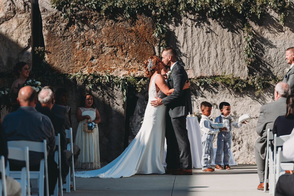 bride and groom share first kiss in front of large slabs of stone covered in ivy at a woolery mill wedding