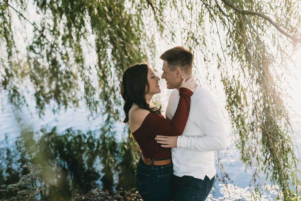 man and woman hug under weeping willow tree at their coxhall gardens engagement session