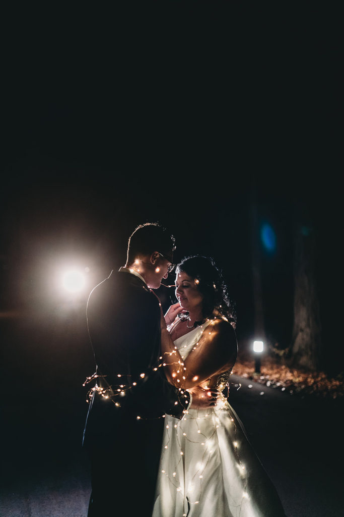 flash pop behind bride and groom during intimate night shot photos while wrapped in fairy lights