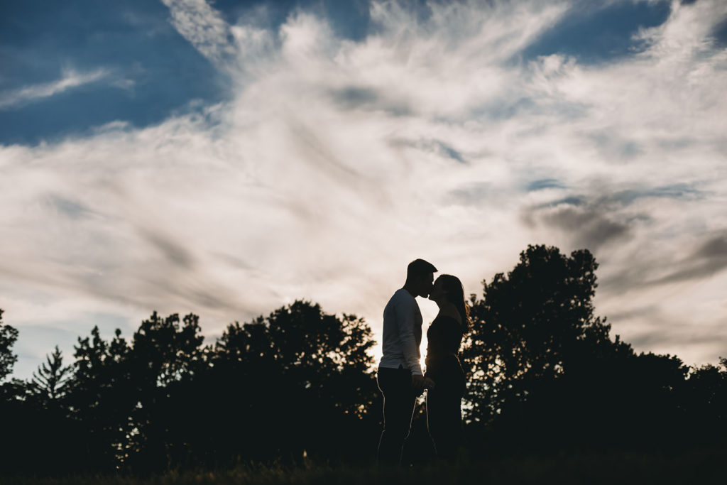silhouette of man and woman kissing in front of cloudy sky at their coxhall gardens engagement session