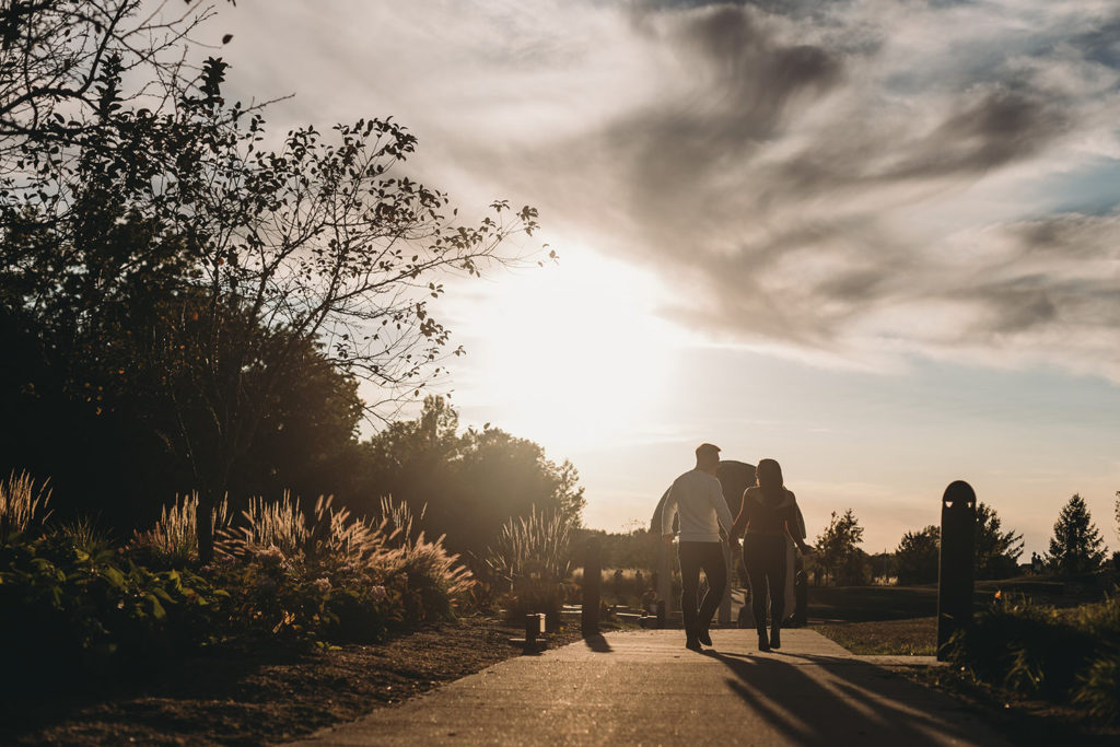 man and woman walk hand in hand toward setting sun at their coxhall gardens engagement session