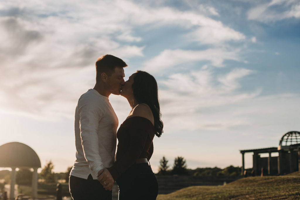 man and woman kiss in front of cloudy sky with lots of white at their coxhall gardens engagement session