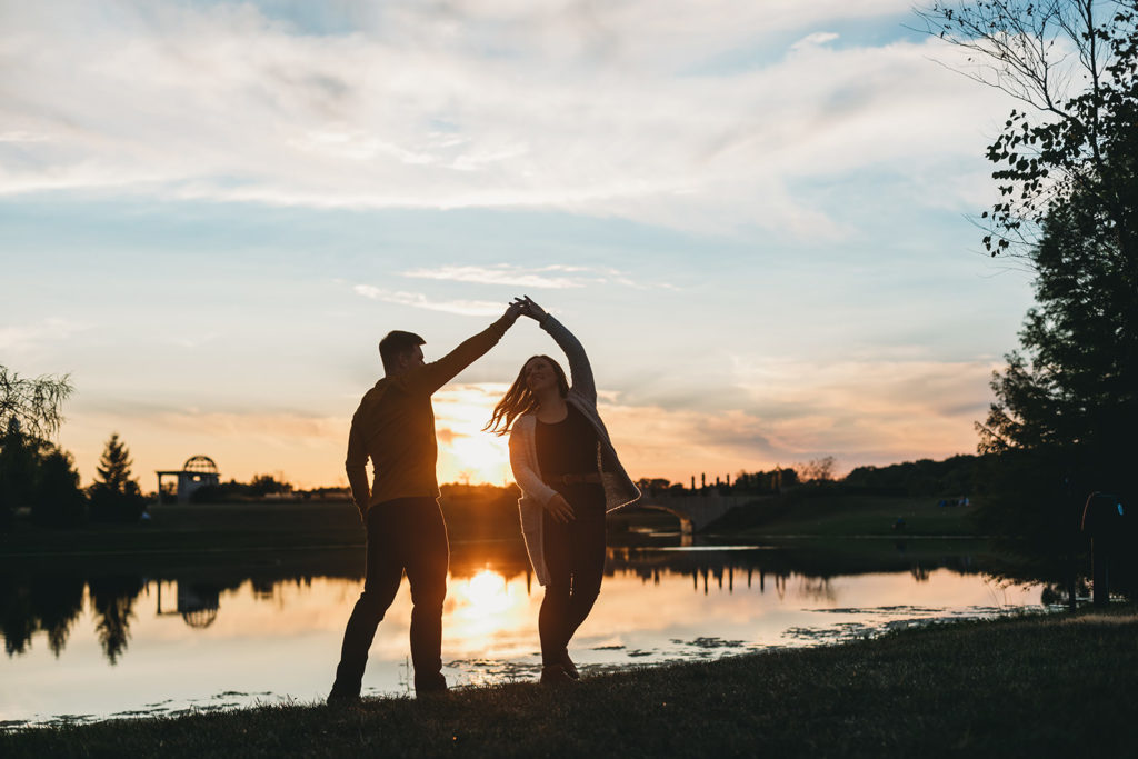 man spins woman while silhouetted in front of pond at sunset at their coxhall gardens engagement session