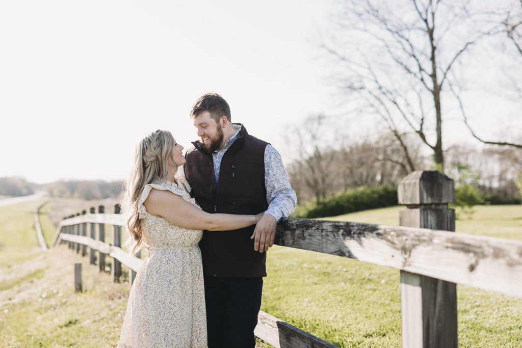 man leans against fence while woman hugs him during their Noblesville park engagement session