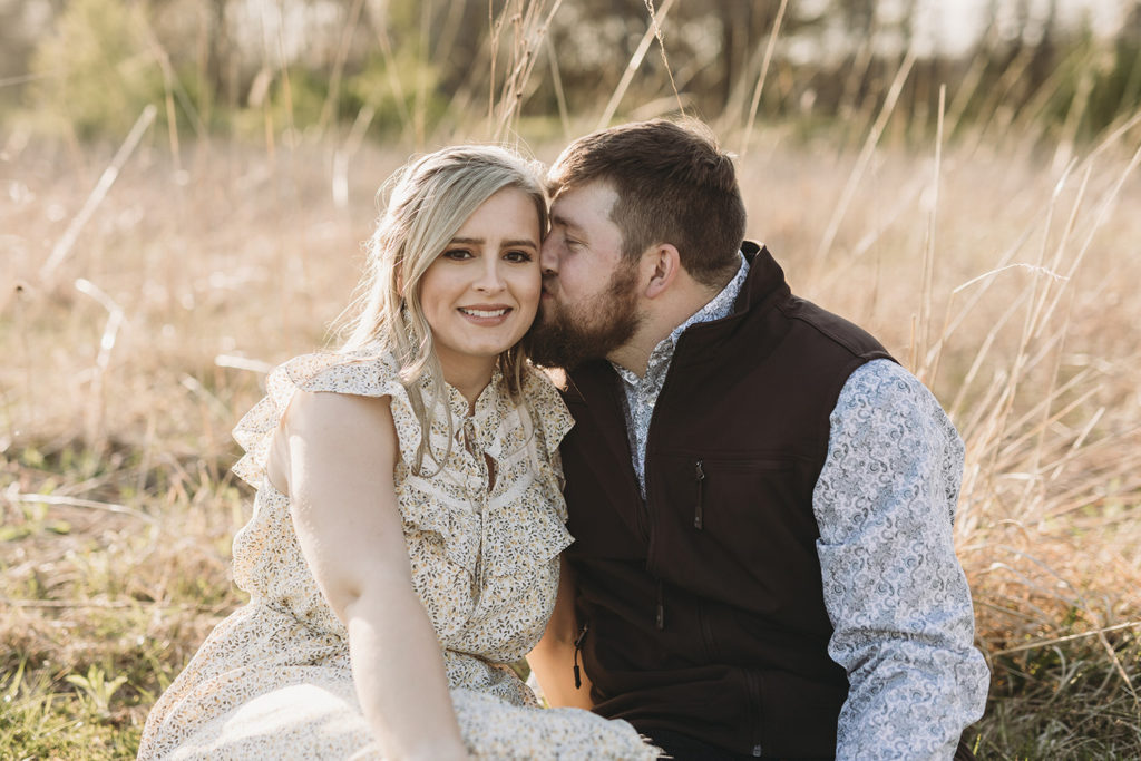 man in vest whispers into blonde woman's ear while sitting in field of tall grass during their Noblesville park engagement session