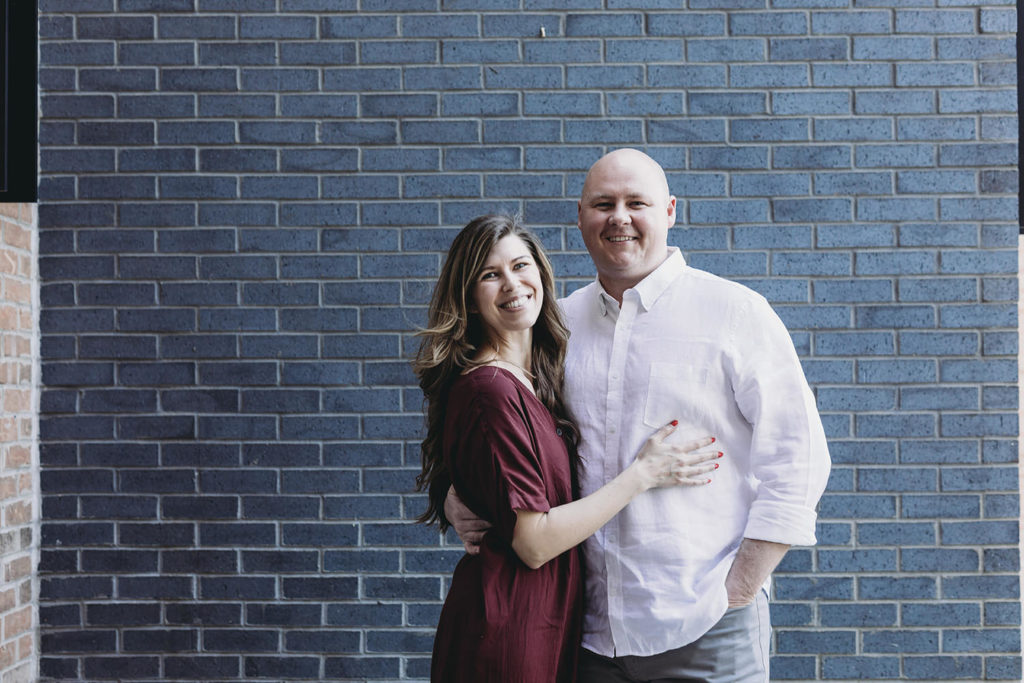 man and woman in front of blue brick wall smiling and hugging during their Fountain Square Engagement Photos