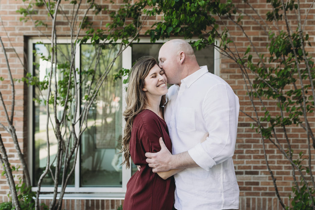 man kisses woman in front of foliage covered brick wall during their Fountain Square Engagement Photos