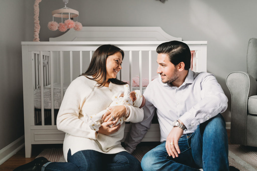 mom holds baby with husband in front of white crib captured by their Indy Newborn Photographer