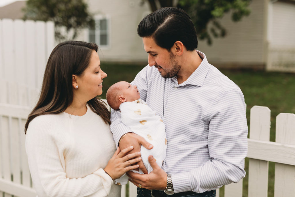 mom and dad hold baby outside by white picket fence captured by their Indy Newborn Photographer