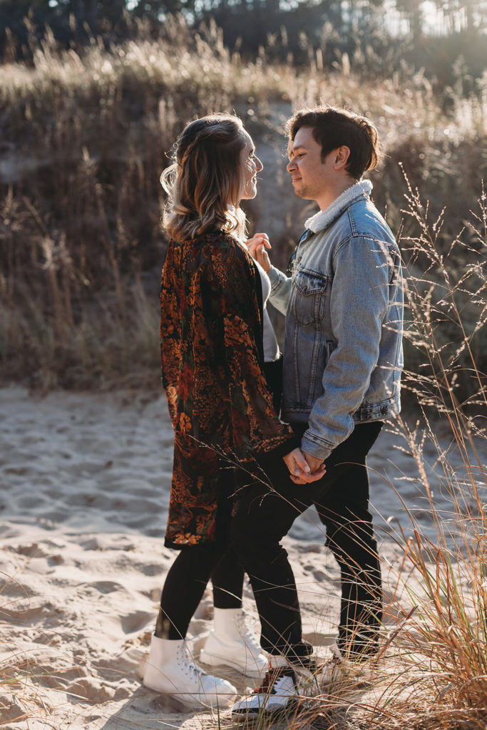 blonde girl in sweater dances with black haired man in jean jacket during their Indiana Dunes engagement photos