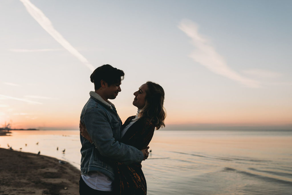 young couple hug at sunset in moody photo on the shore of Lake Michigan in Indiana