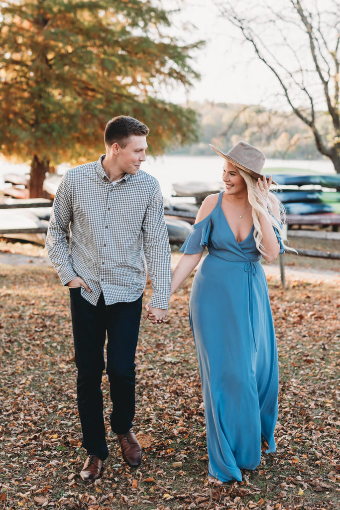 blonde woman in blue dress with wide brimmed hat holds hands with man in gray shirt in front of canoes during their eagle creek photo session