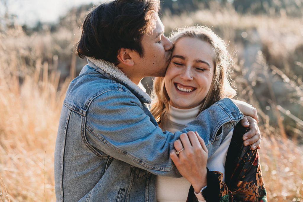 man in jean jacket kisses woman in sweater jacket during their Indiana Dunes engagement photos