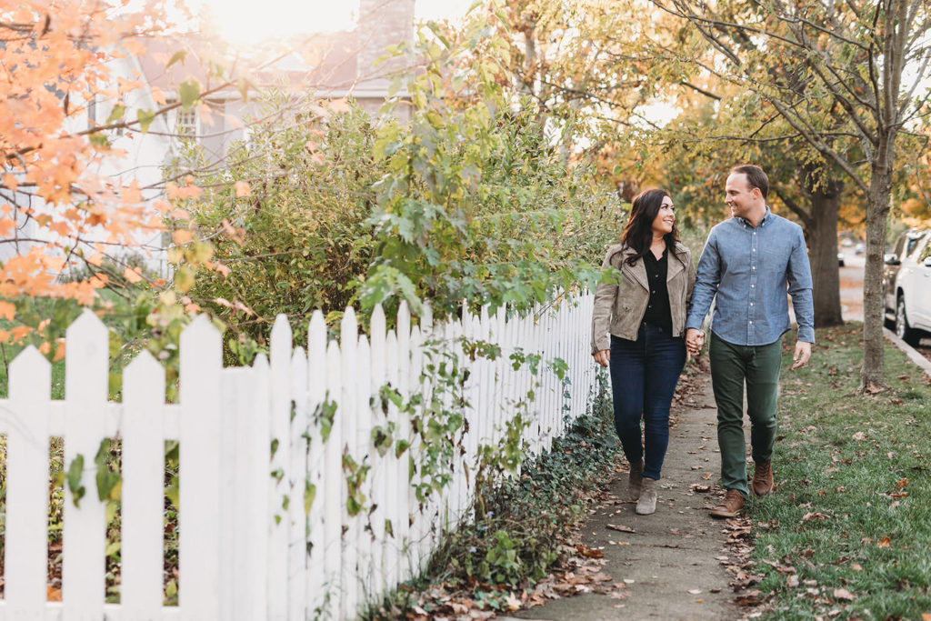 couple holds hands and walks past white picket fence and trees with fall colors during their downtown zionsville engagement session