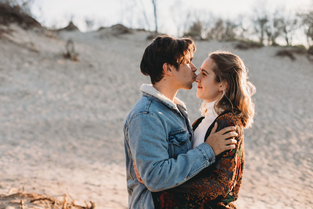 young man in jean jacket kisses blonde woman on the nose during their Indiana Dunes engagement photos