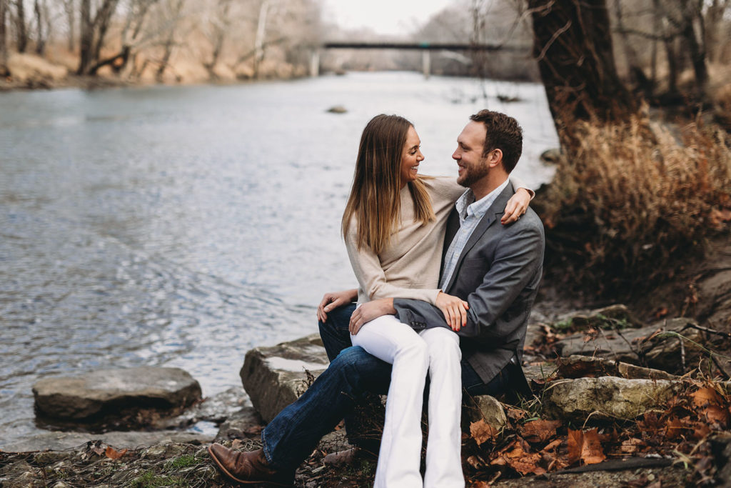 young engaged couple sit on banks of white river captured by their Noblesville Photographer
