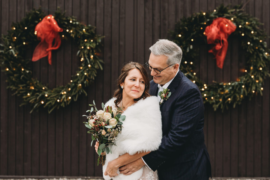 groom hugs bride from behind while they stand in front of christmas wreaths during their traders point creamery elopement