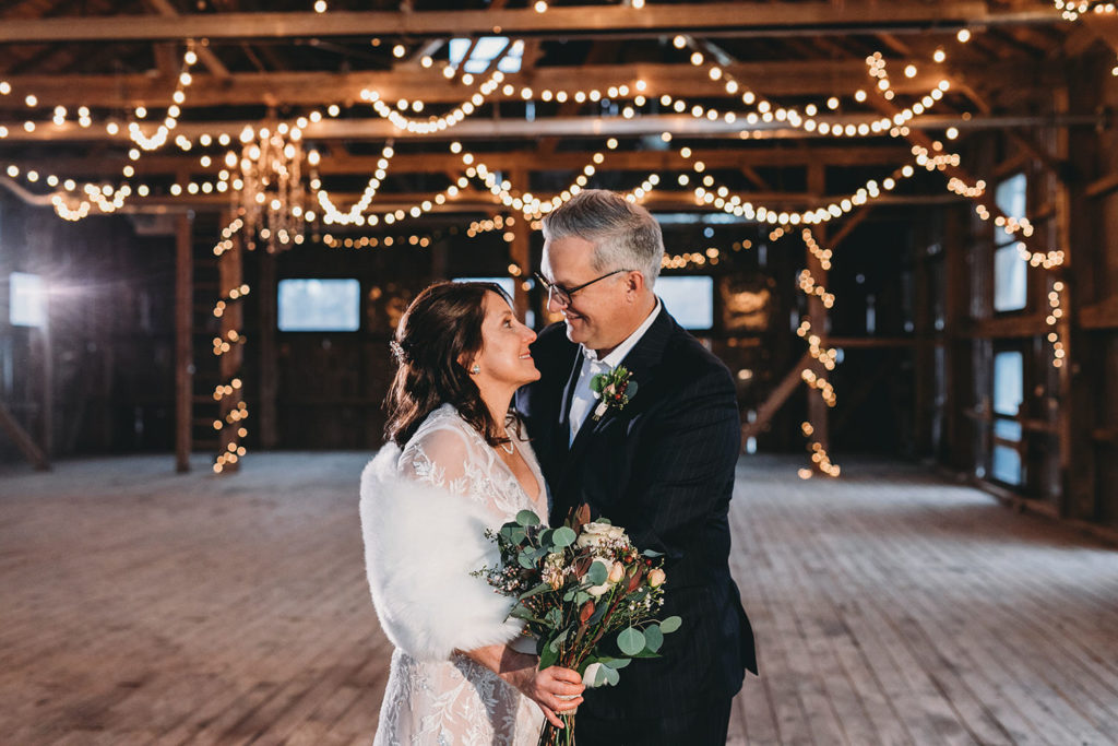 bride and groom look at each other and smile during their traders point creamery elopement in a barn with string lights