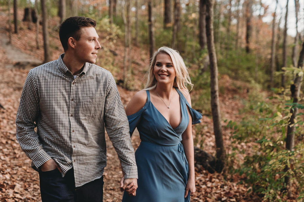blonde woman in blue dress and man in gray shirt hike through woods during their eagle creek photo session